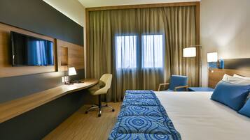 Habitación de hotel con cama, escritorio, silla, televisor y ventana con cortinas.