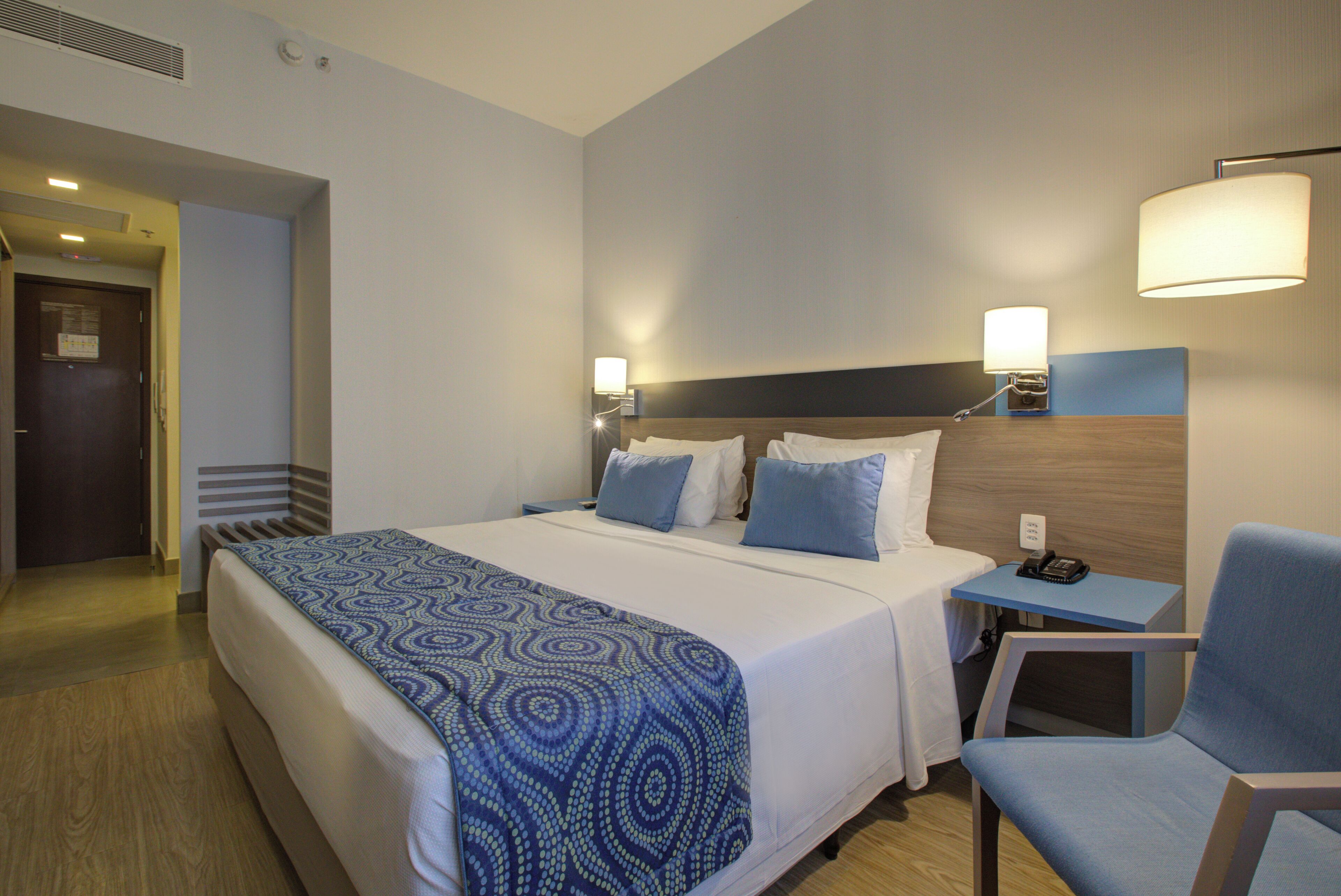 Una camera d'albergo con un letto grande, un comodino con una lampada, una sedia e una porta che dà su un corridoio.