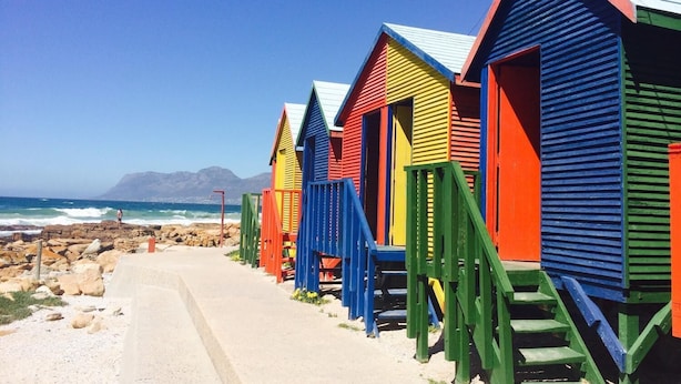 A row of colorful beach huts with steps leading up to them, situated on a sandy beach with a rocky shoreline and a mountain in the background. - Harbour Place (Cape Town)