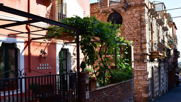 Una calle estrecha con un edificio de piedra, una reja de hierro forjado y un letrero que dice 'Casa del Ginnasio'.