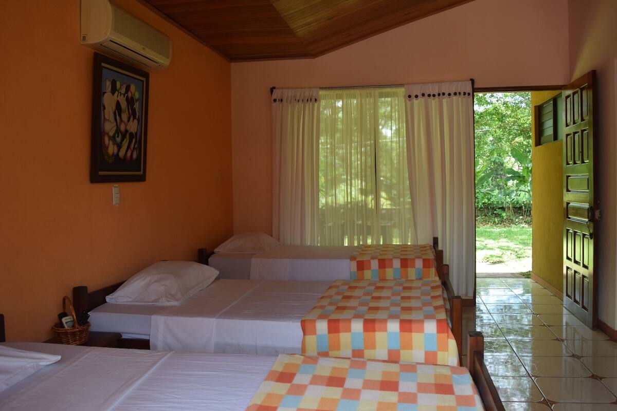 A hotel room with three beds, a painting on the wall, and a view of greenery outside.