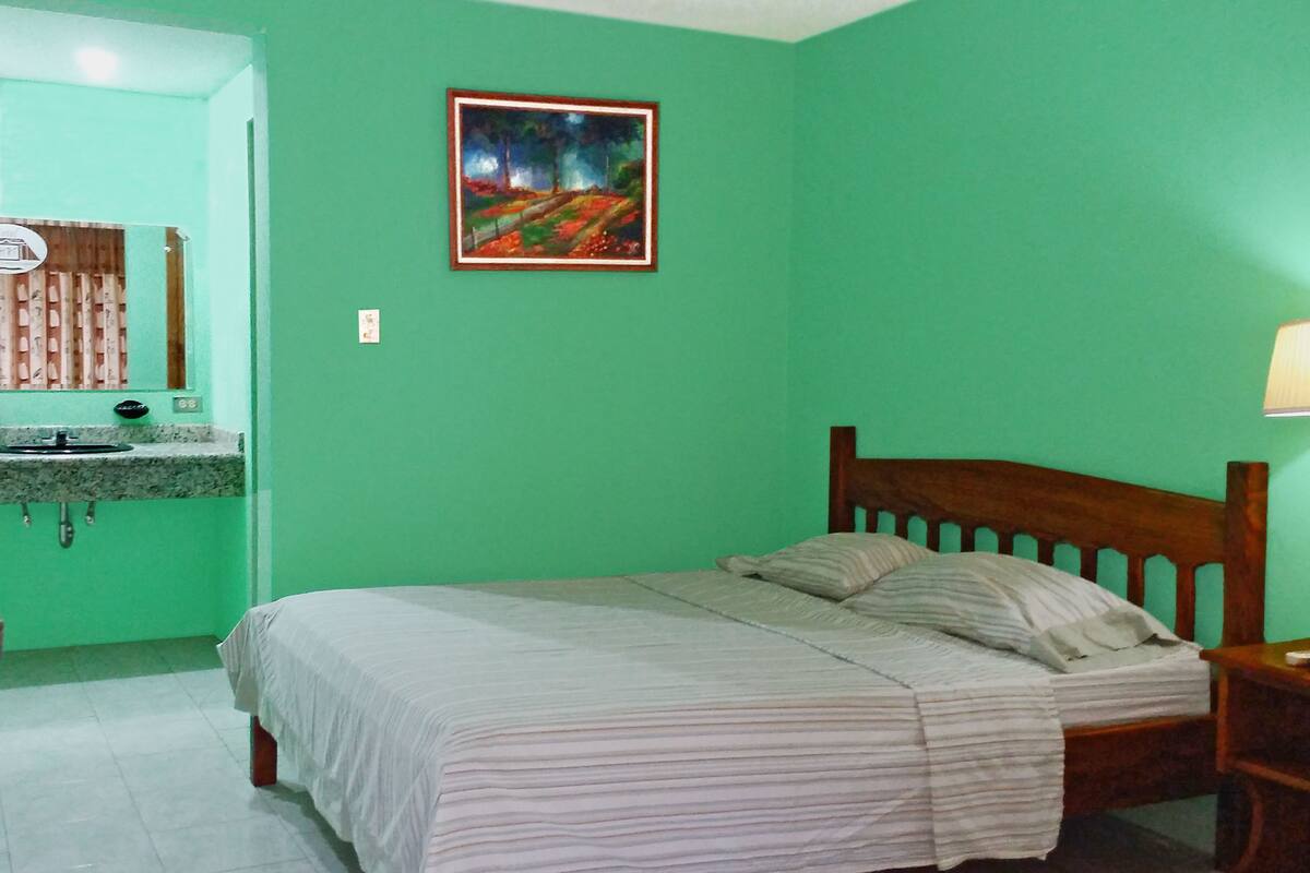 A bedroom with a bed, a nightstand, a wooden headboard, a green wall, a painting, a window with blinds, and a bathroom with a sink and mirror.