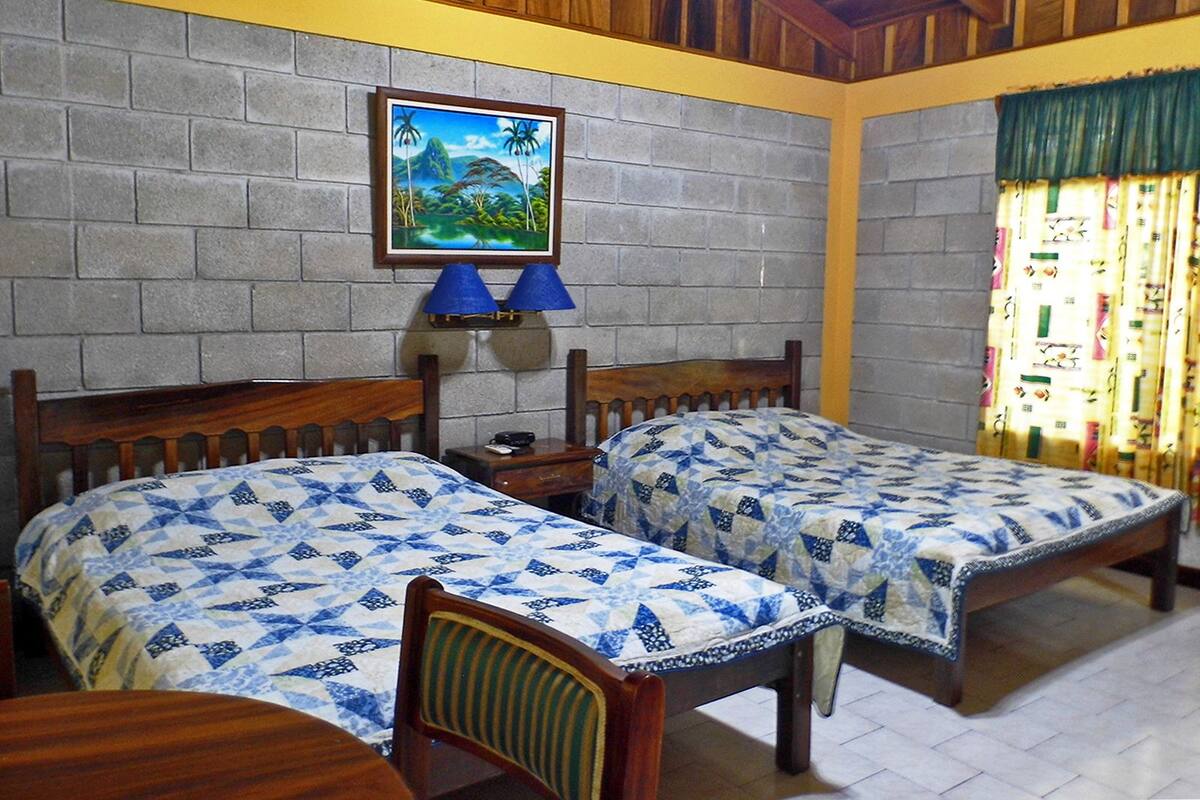 A room with two wooden beds, a small table, a chair, a wall-mounted lamp, and a painting.