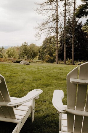 Relax in serene surroundings with two Adirondack chairs on a lush lawn overlooking a natural backdrop of trees and a rock formation.