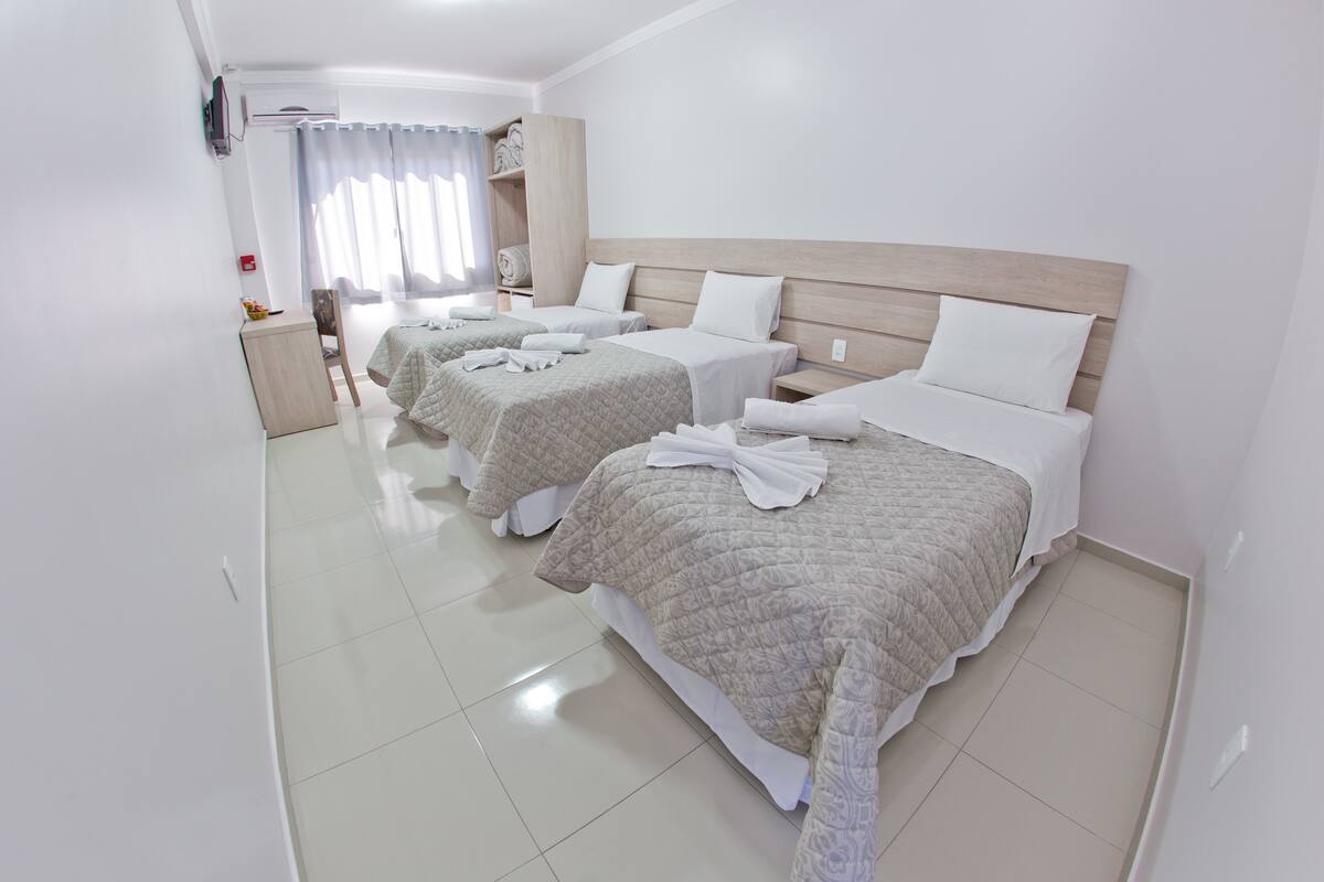 A hotel room with three beds, each with a white headboard and a quilted bedspread. There is a small table with a chair, a nightstand, and a TV mounted on the wall.