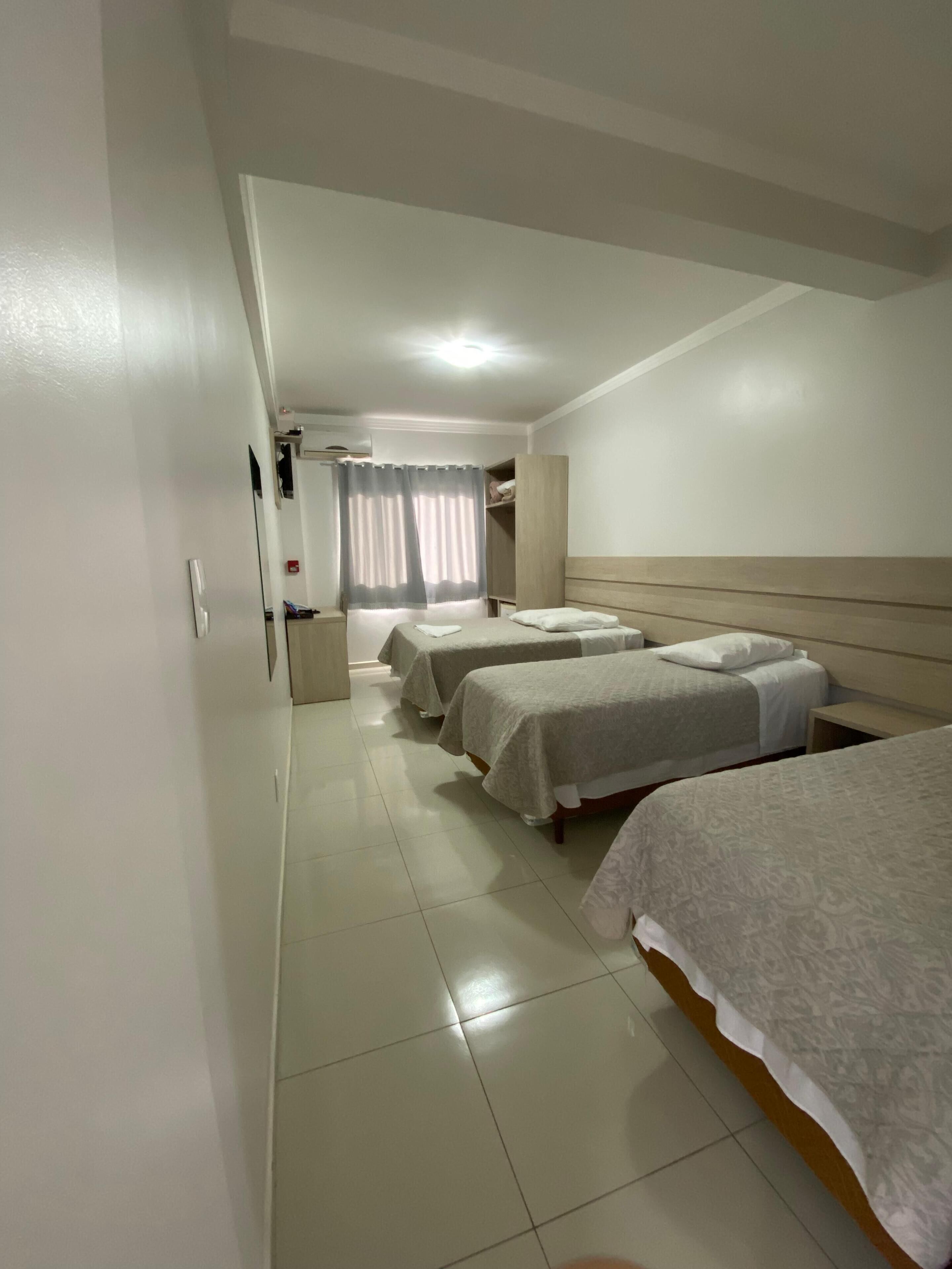 A hotel room with four beds arranged in a row, a large window with curtains, and a tiled floor.