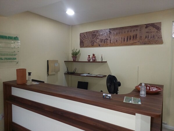 A hotel reception desk with a wooden counter, a signboard with hotel information, a wall-mounted plant, and a decorative mural.
