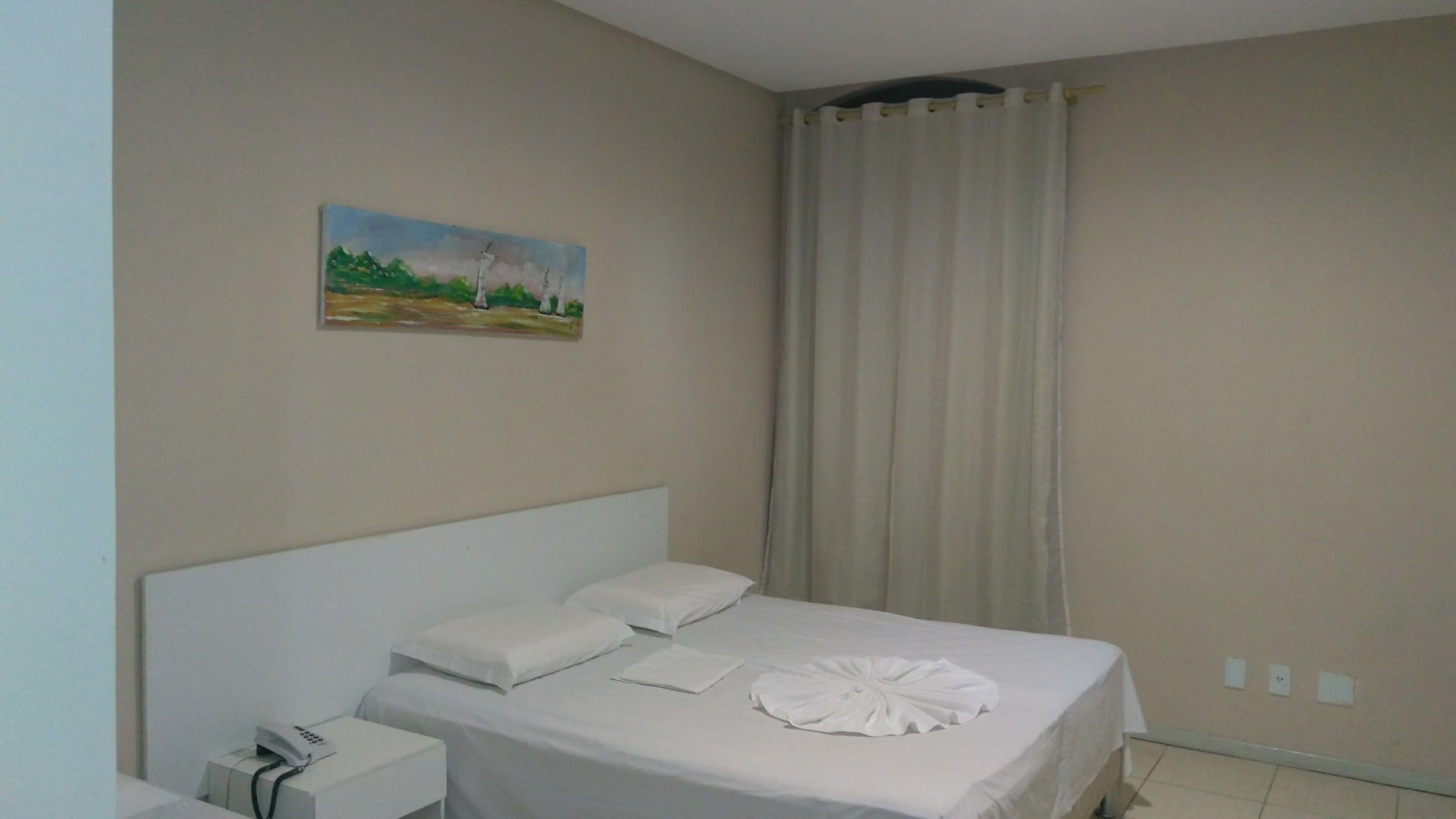 Ein ordentlich bezogenes Bett mit weißen Leinen, ein Bild an der Wand und ein Vorhang, der ein Fenster verdeckt.