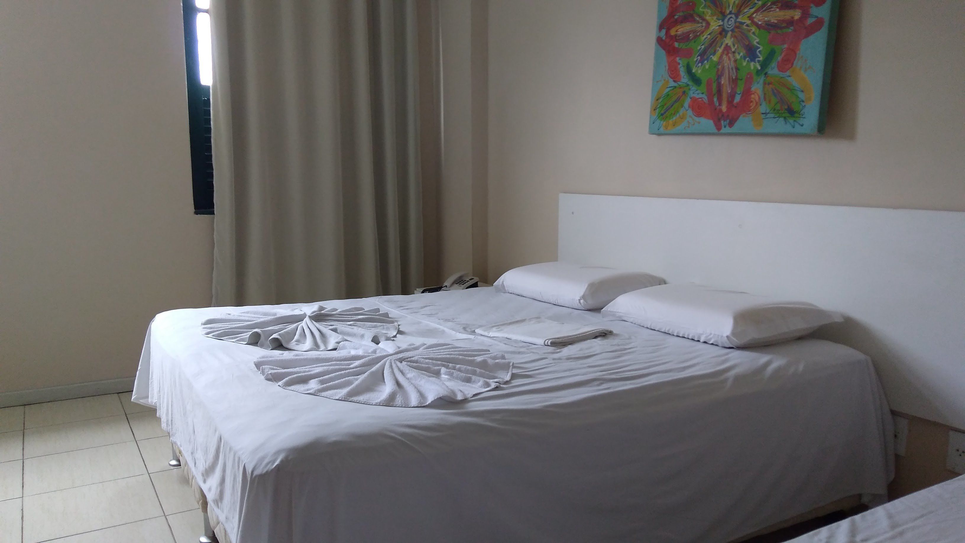 A neatly made bed with white linens and a decorative towel arrangement. A telephone and a painting are visible on the wall.