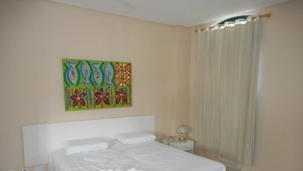 A neatly made bed in a hotel room with a plain wall and a window with curtains. - Cachoeira Apart Hotel (Cachoeira)