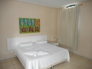A neatly made bed in a hotel room with a plain wall and a window with curtains.