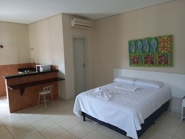 Quarto de hotel com cama, secretária pequena com micro-ondas, uma cadeira e porta.