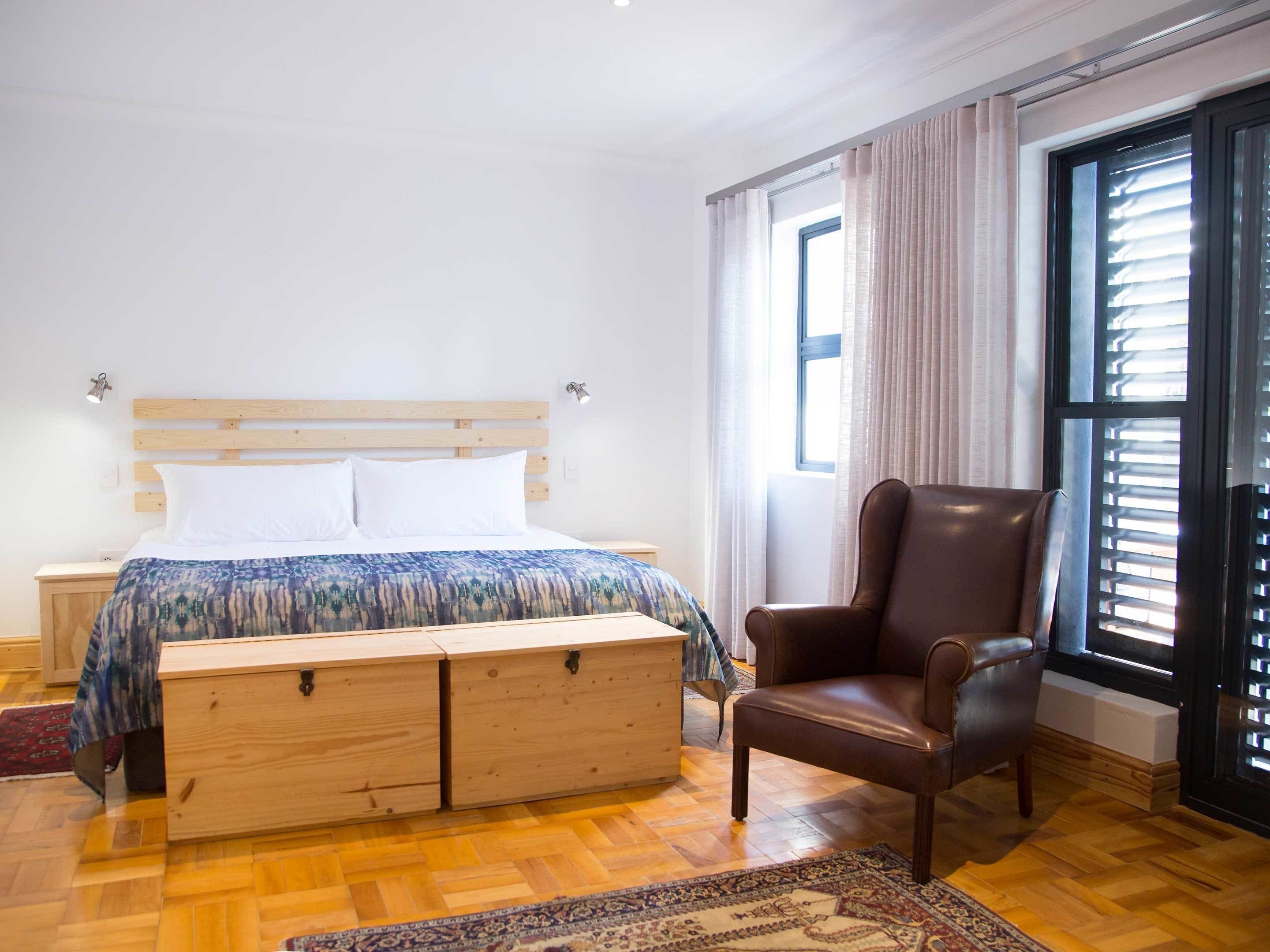 Un dormitorio con una cama de madera, un sillón de cuero, un aparador de madera y un ventanal con persianas.
