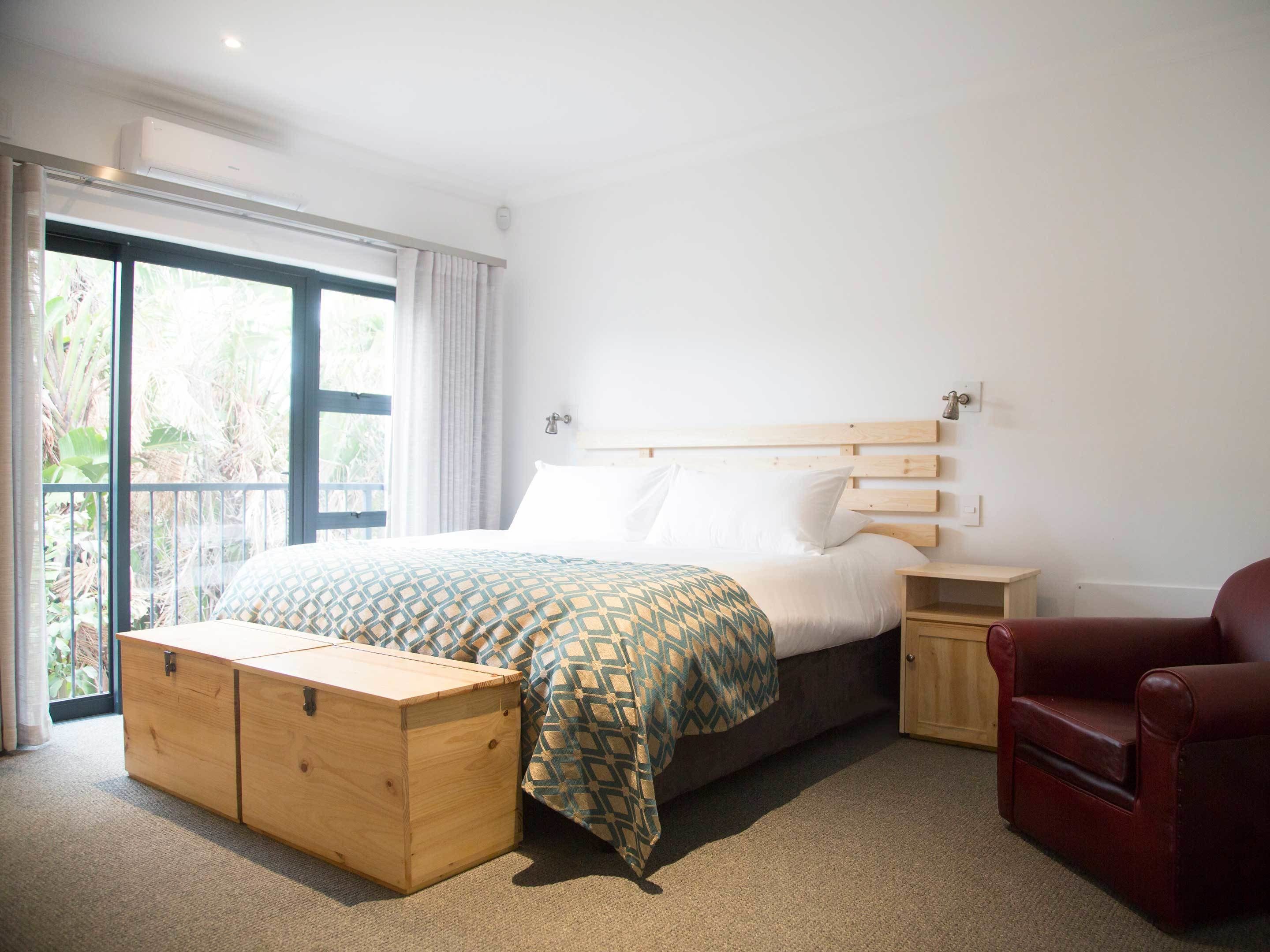 Un dormitorio con una cama, un banco de madera, una silla roja y un ventanal corredizo que da a un balcón.