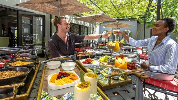 An outdoor buffet with various food items and drinks, set up under umbrellas with a view of a building in the background. - La Fontaine (Franschhoek)
