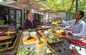 Un bufé al aire libre con diferentes alimentos y bebidas, dispuesto bajo sombrillas y con vista a un edificio al fondo.