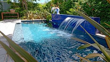 A swimming pool with a waterfall feature, surrounded by lush greenery and a wooden bench.