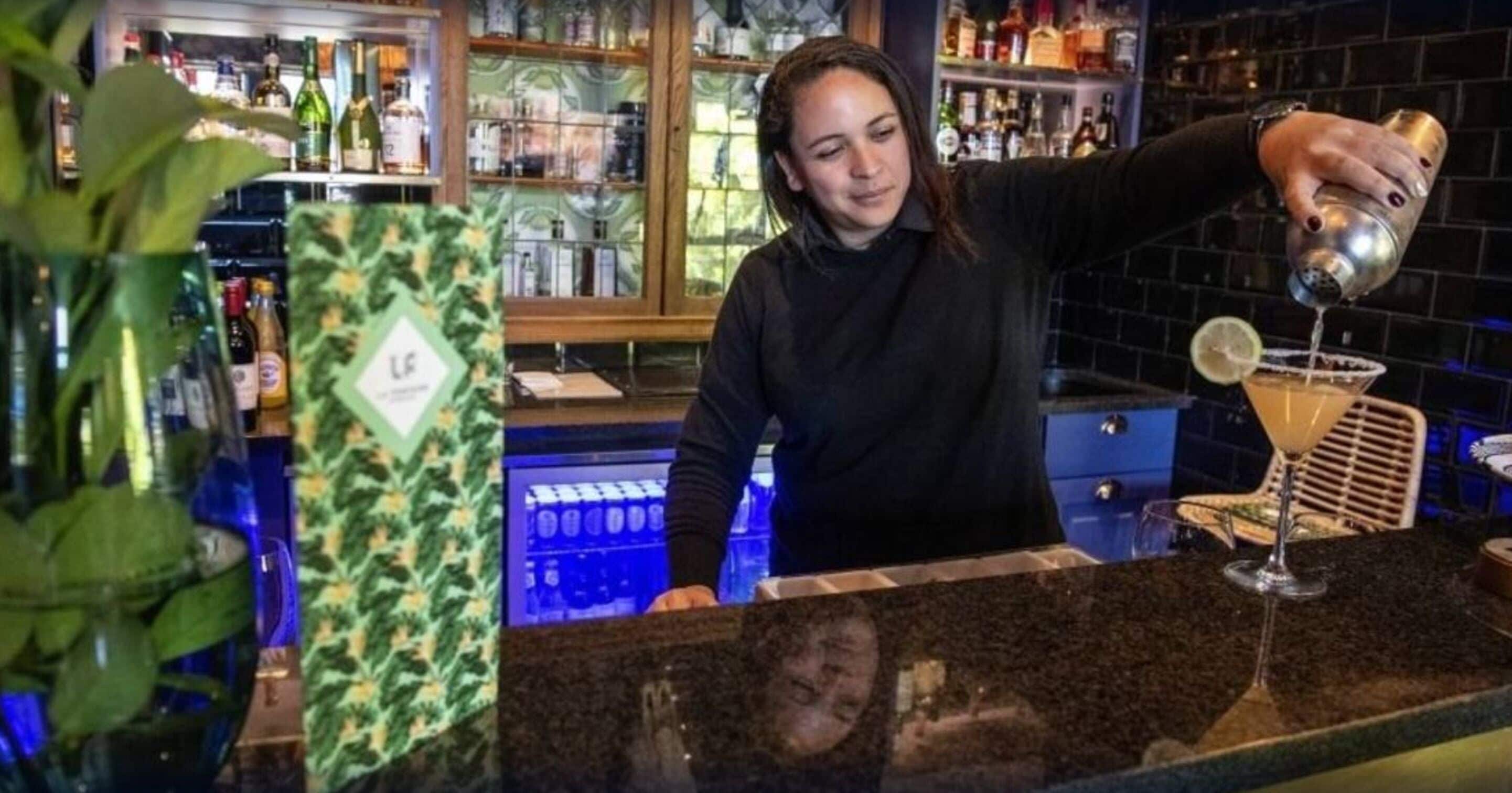 Uma área de bar com um bartender preparando uma bebida. Há prateleiras com garrafas e uma planta ao primeiro plano.
