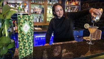 Uma área de bar com um bartender preparando uma bebida. Há prateleiras com garrafas e uma planta ao primeiro plano.