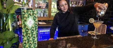 Un coin bar où un barman prépare un cocktail. On aperçoit des étagères remplies de bouteilles et une plante au premier plan.