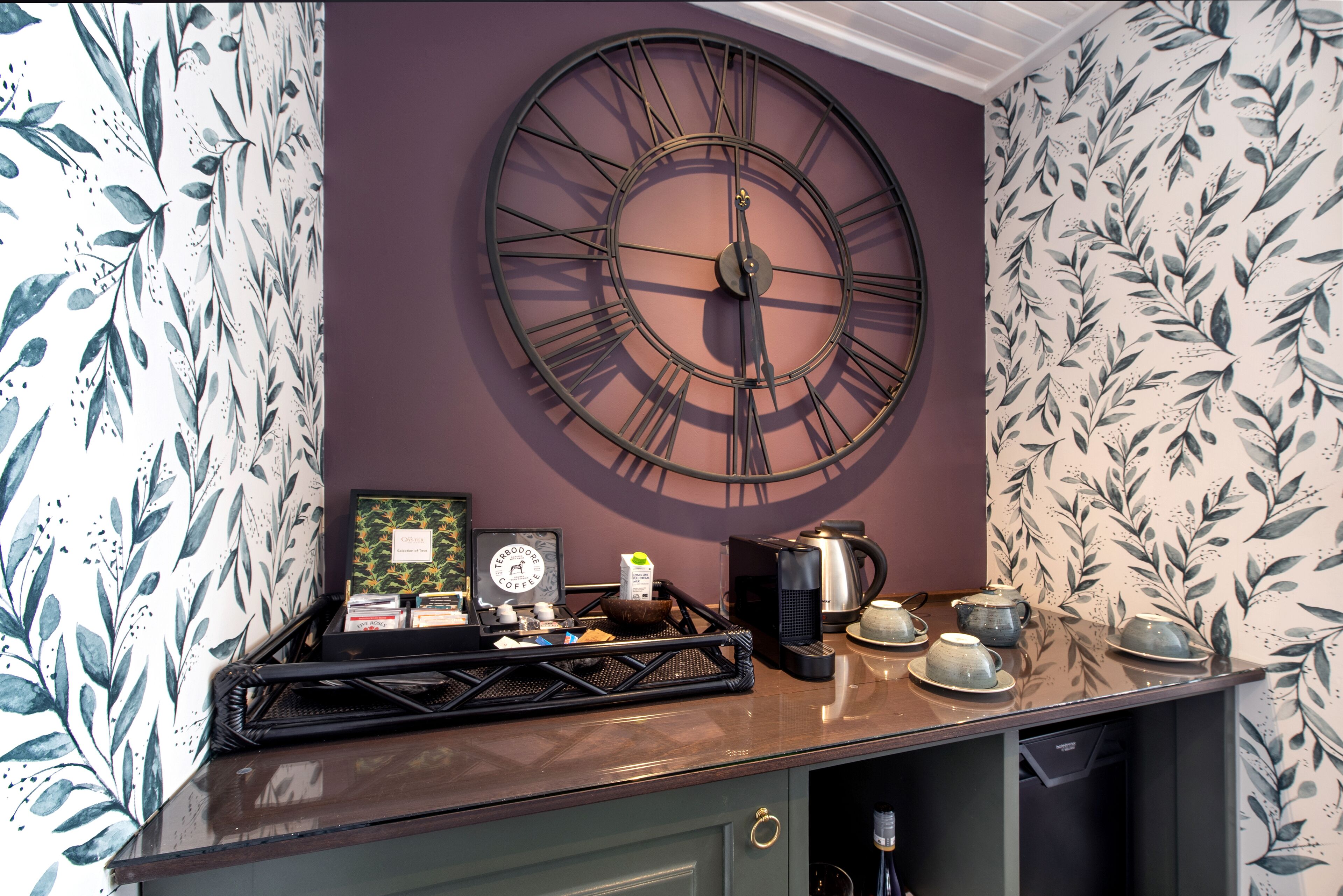 Una cocina con un gran reloj de pared, una cafetera y una bandeja con tazas y una tetera.