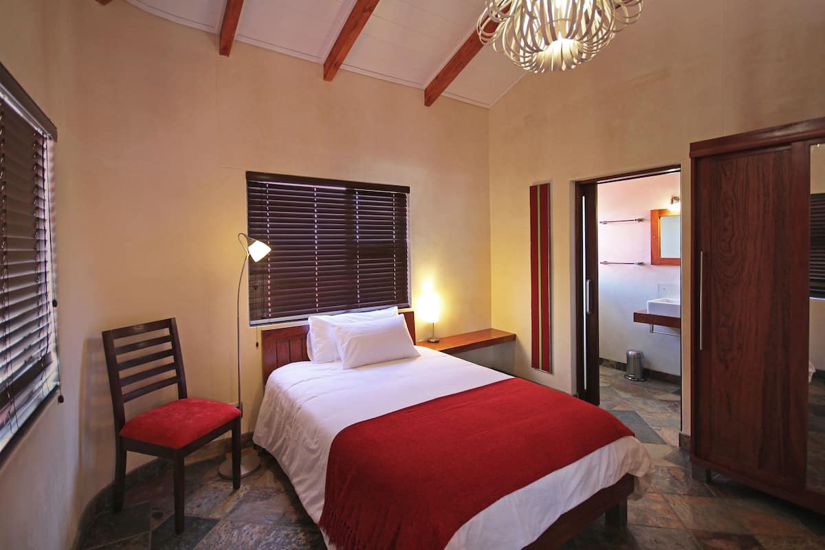 Quarto com cama, cadeira, mesa de cabeceira, abajur e janela com persianas.