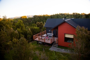 A red cabin with a wooden deck and a single window, surrounded by dense forest.