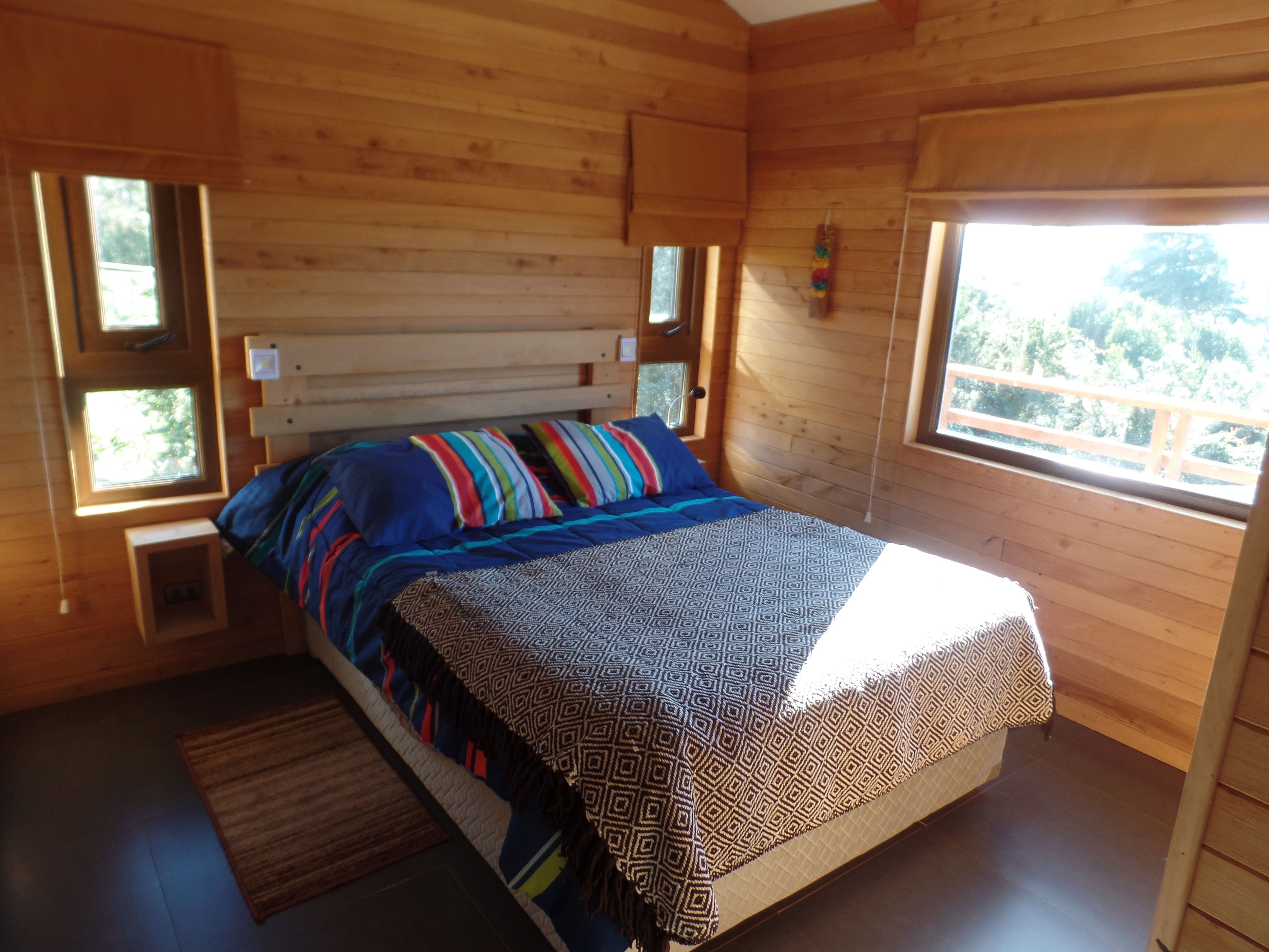 Ein Schlafzimmer einer Holzhütte mit einem Bett, zwei Fenstern und Blick auf den Wald.