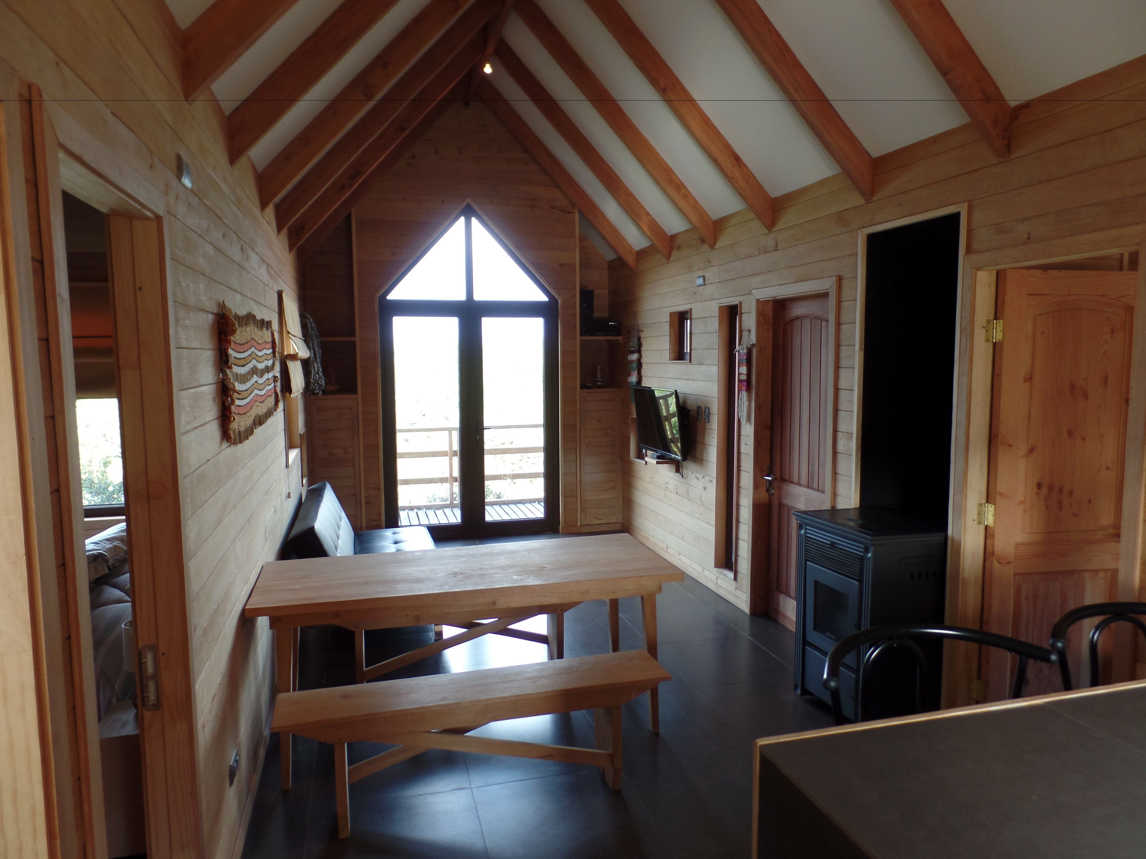 A cozy cabin interior with a wooden dining table, benches, a fireplace, and a large window providing natural light.