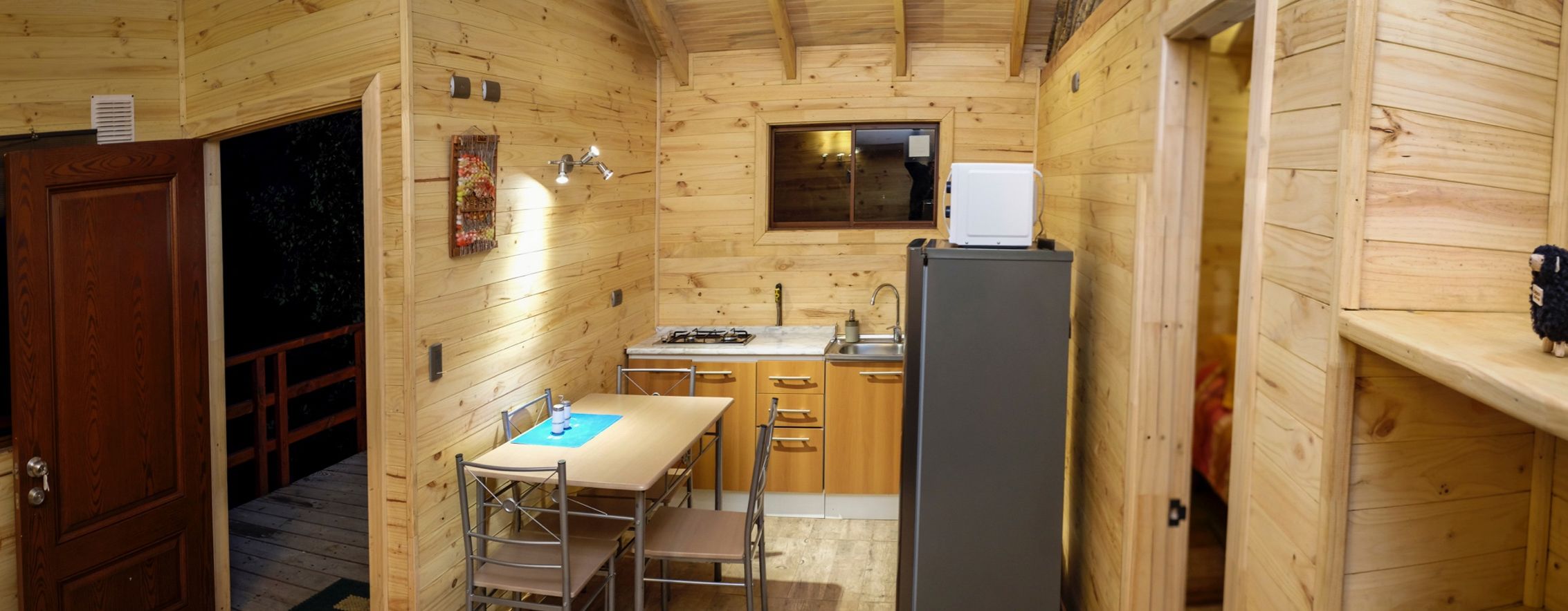 A compact wooden cabin with a kitchenette, a dining area with metal chairs, and a small window.