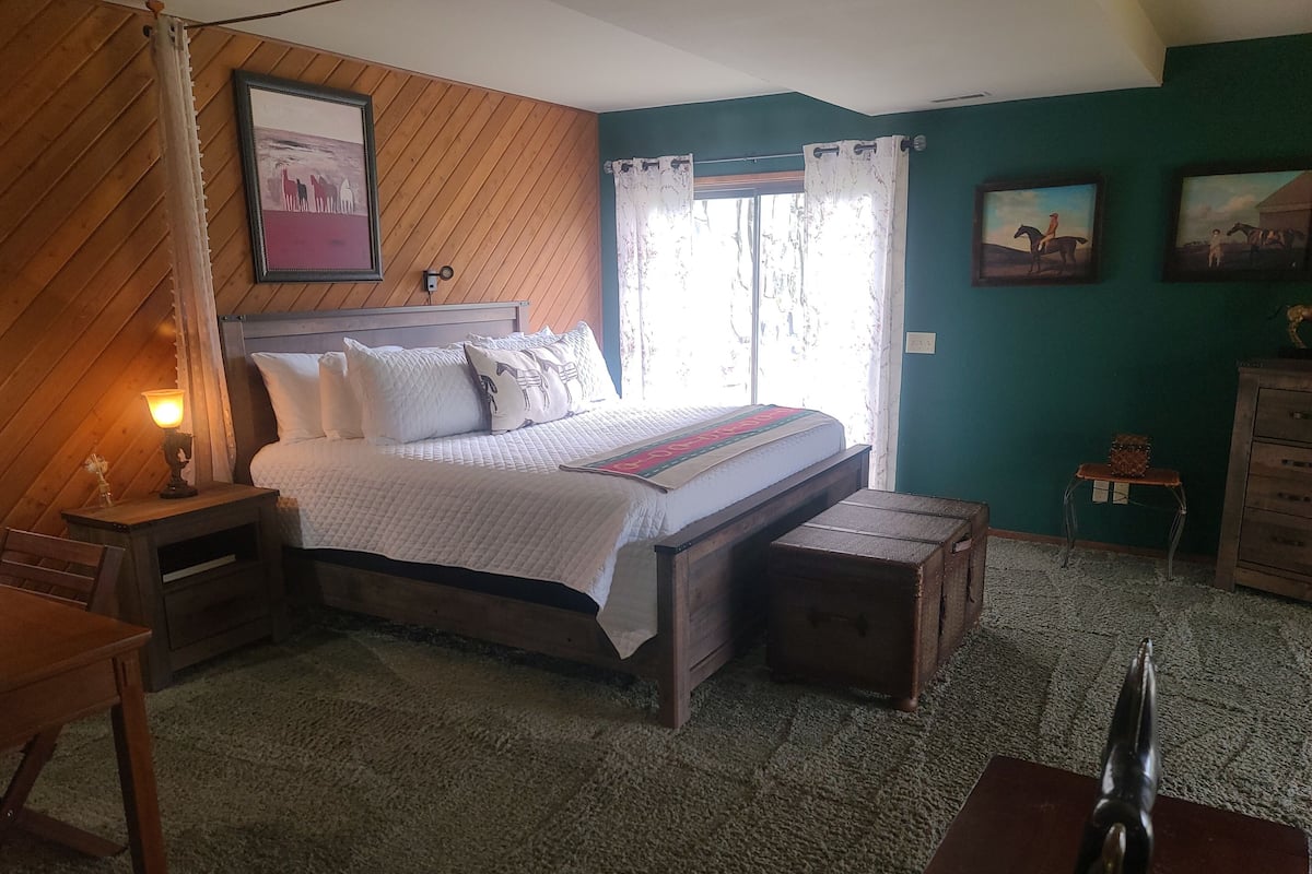Un dormitorio con cama, cabecera de madera, mesita de noche con lámpara, una pequeña mesa, maleta y cómoda.