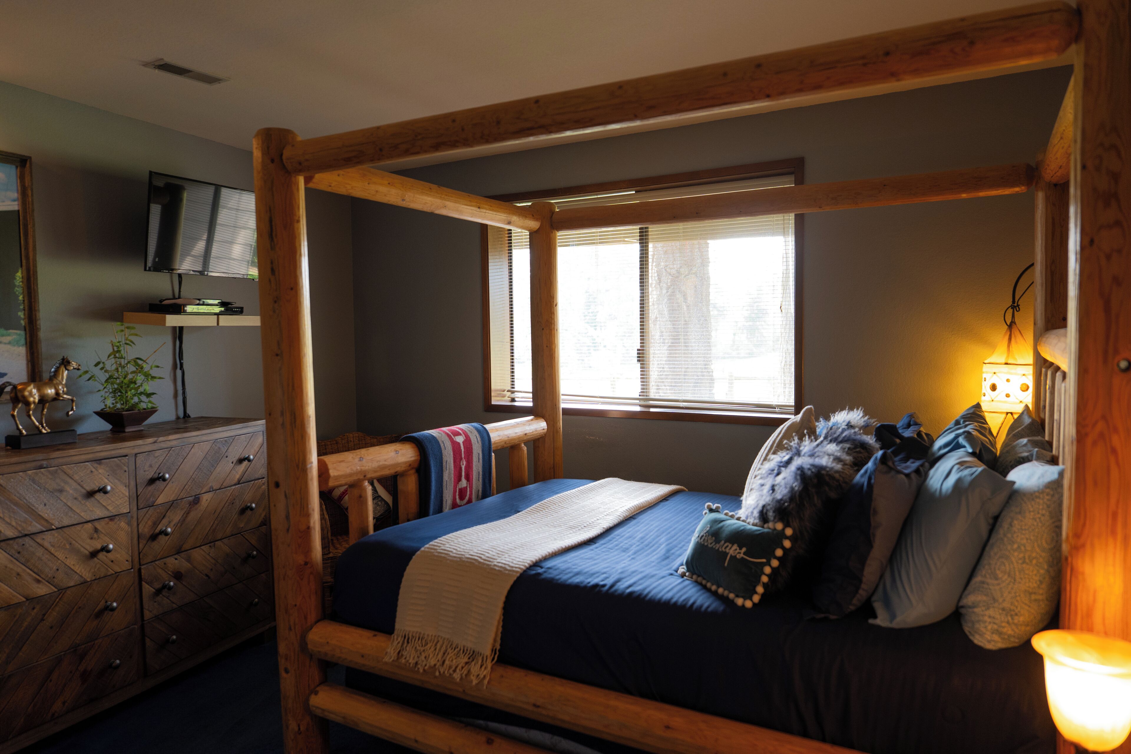 A room with a wooden four-poster bed, a television on a wooden cabinet, and a window with blinds.