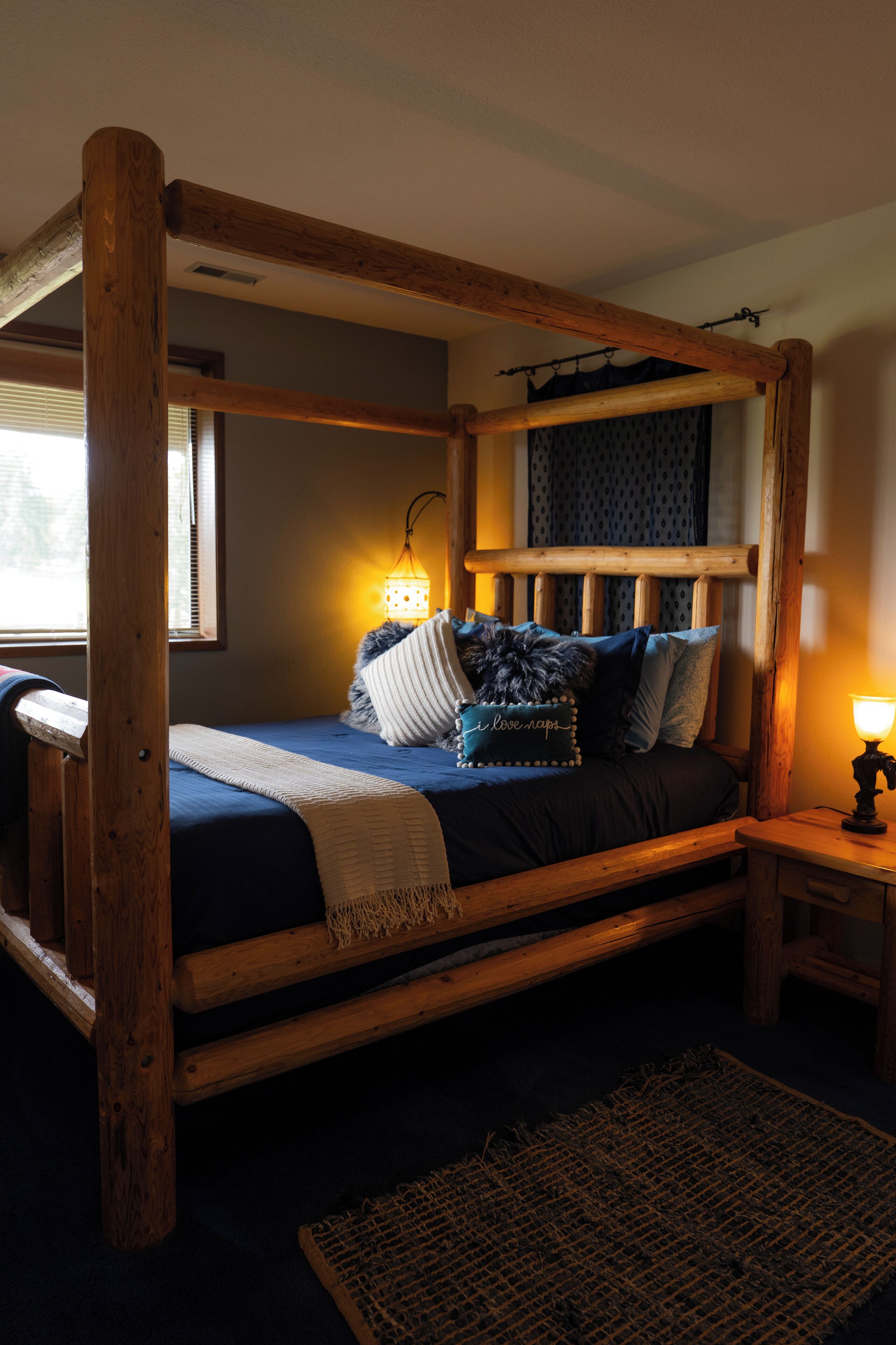 A wooden four-poster bed with a dark blue bedspread and pillows, a small bedside table with a lamp, and a window with blinds.
