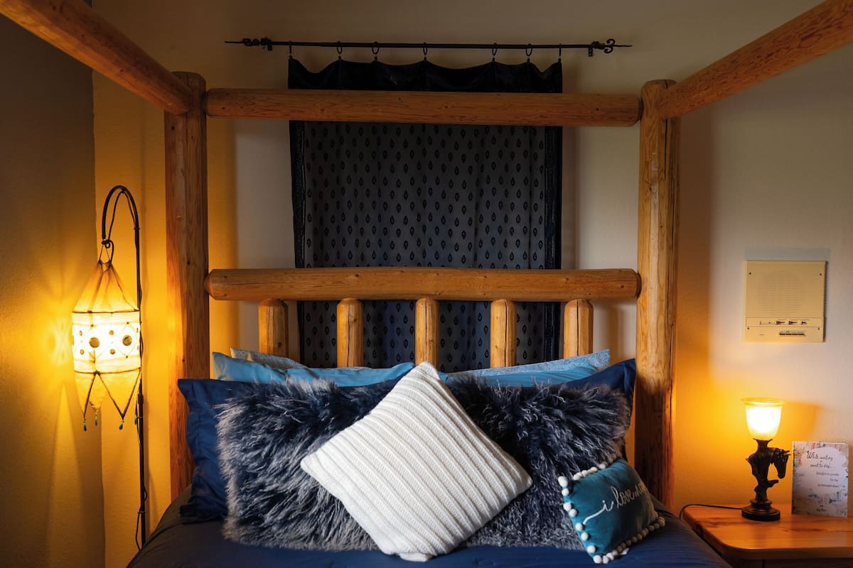 Una cama con estructura de madera y cabecera, una lámpara fijada a la pared y una mesita de noche con lámpara.