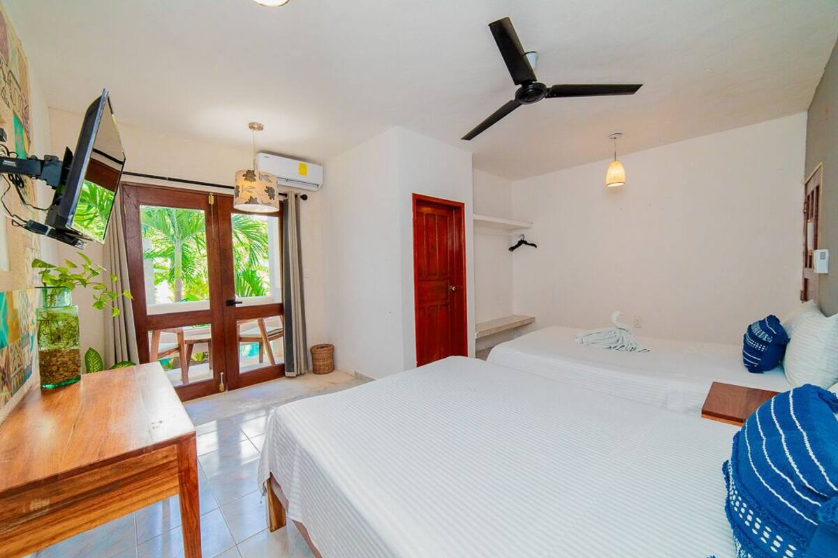 A bedroom with two beds, a desk, a ceiling fan, and a window with a view of palm trees.
