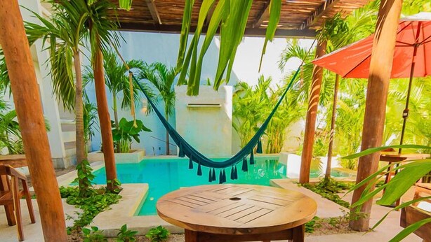 Une table et des chaises en bois, placées sous une pergola en bois, avec un hamac, le tout entouré de palmiers et d’une piscine.