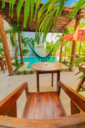 A wooden table and chairs under a wooden pergola with a hammock, surrounded by palm trees and a swimming pool.