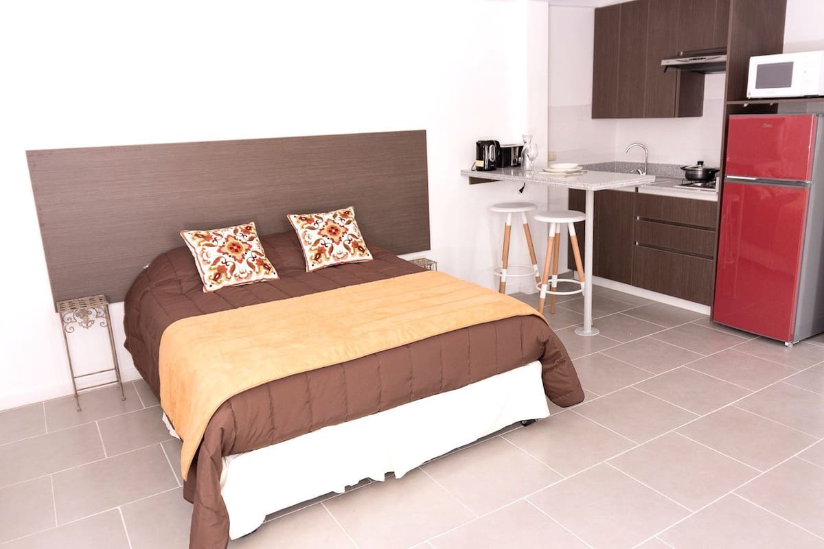 A modern hotel room with a bed, a kitchenette, and a red refrigerator.