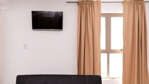 Un salon moderne avec un canapé noir, un téléviseur fixé au mur et des rideaux de couleur beige.