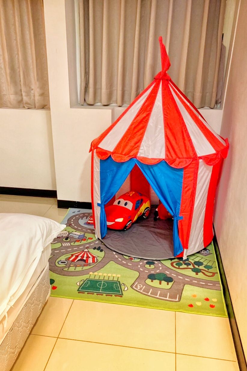 A child's play tent with a toy car inside, a bed with a white cover, and a play mat with a road and car theme.