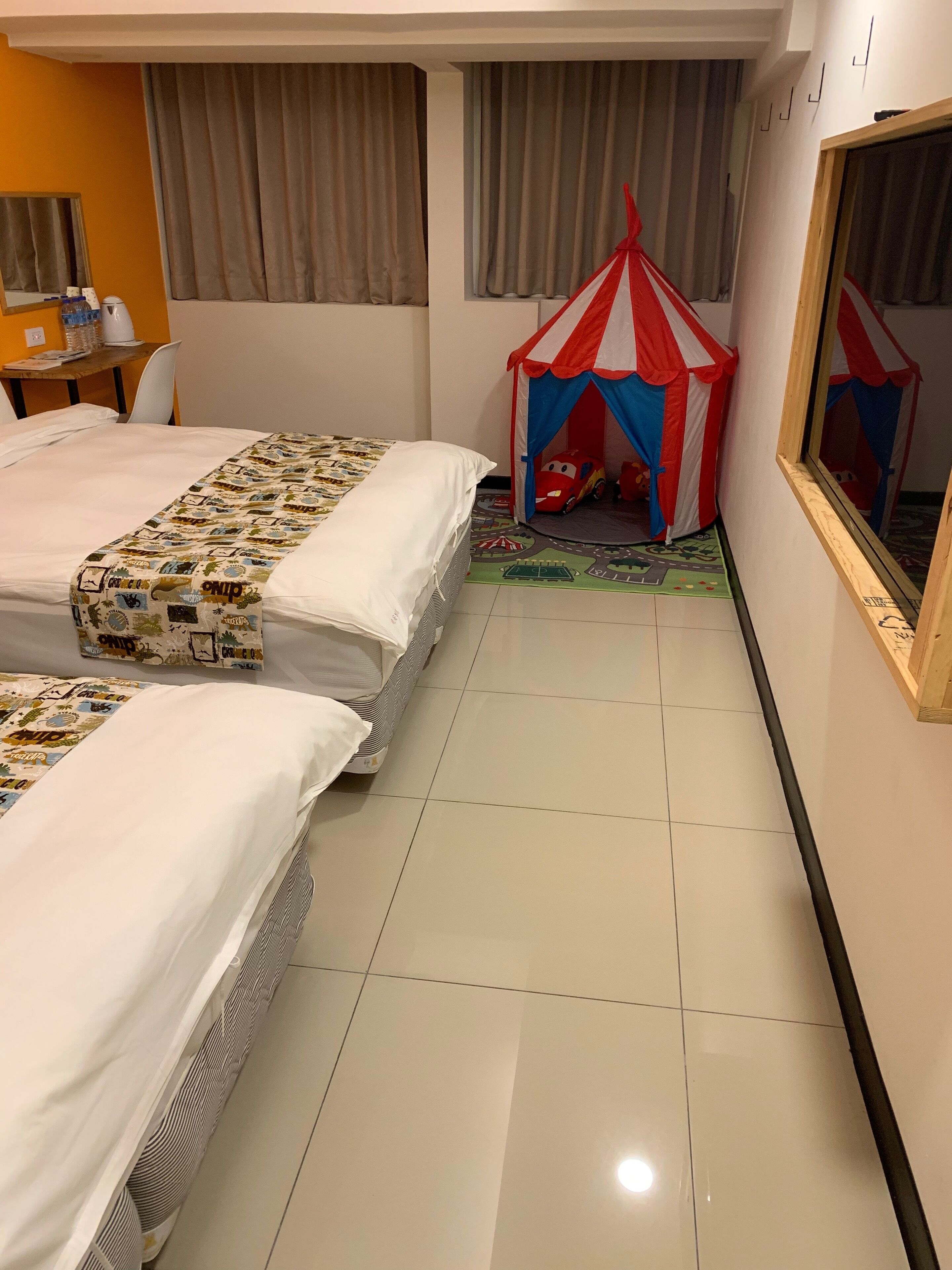 A hotel room with two beds, a small table, a chair, a window, and a red and white tent-like structure.