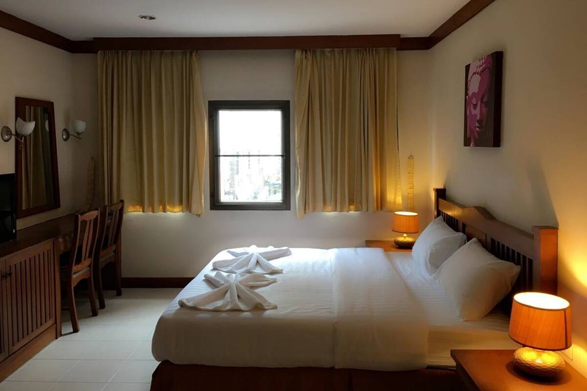 Habitación de hotel con cama, escritorio, silla, ventana con cortinas, lámpara y un cuadro en la pared.