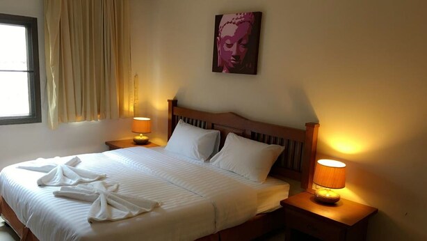 A neatly made bed with white linens and two pillows, flanked by two bedside tables with lamps, in a room with a window and a wall-mounted artwork. - Same Same Guesthouse (Patong)