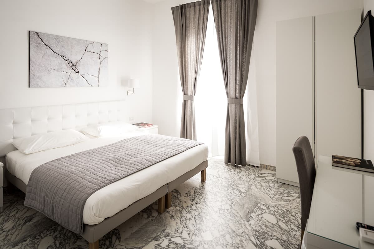 Un dormitorio moderno con suelo de mármol, una cama grande con colcha gris, una mesita de noche con lámpara y un televisor sobre un mueble.