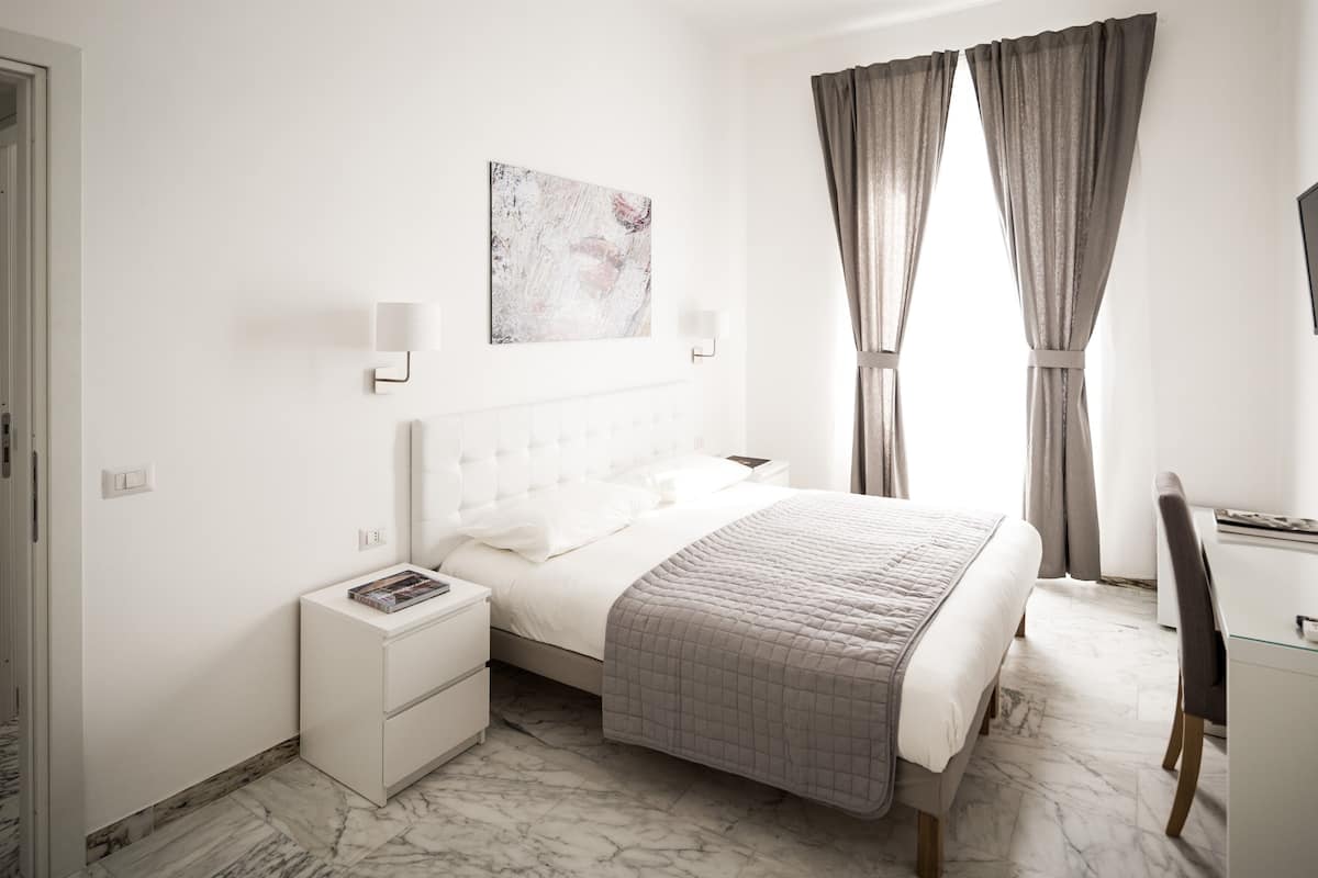 Un dormitorio moderno con suelo de mármol, una cama con colcha acolchada, una mesita de noche con un libro, una lámpara de pared y un cuadro sobre la cama.