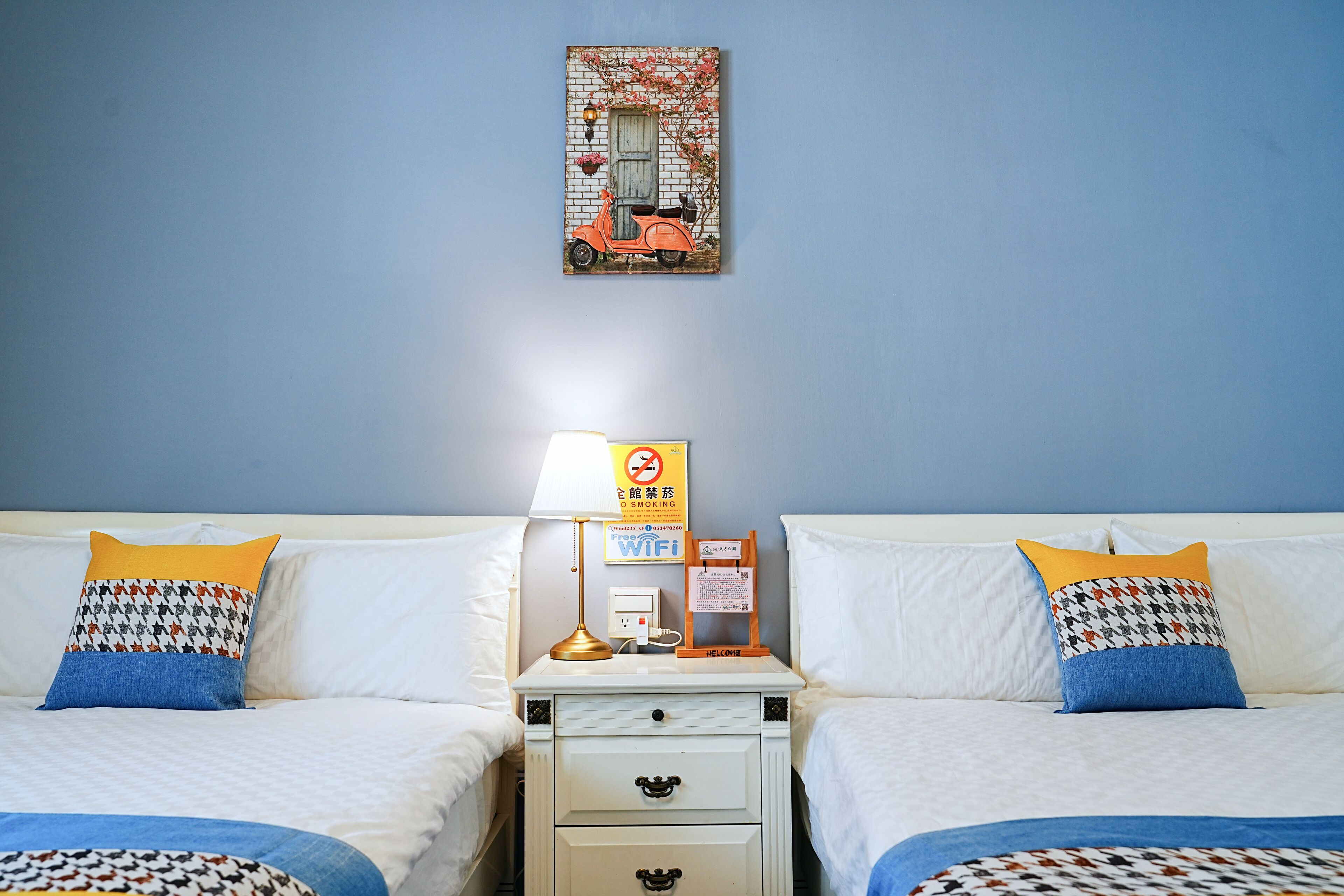Camera d'albergo con due letti, un comodino con una lampada, un cartello a parete e un quadro incorniciato sopra i letti.