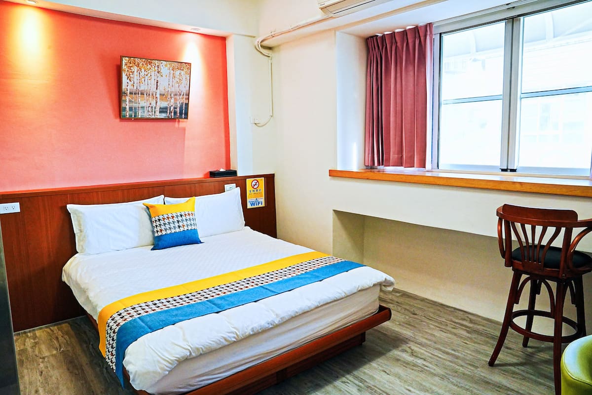 A hotel room with a bed, a chair, a window with curtains, and a wall-mounted artwork.