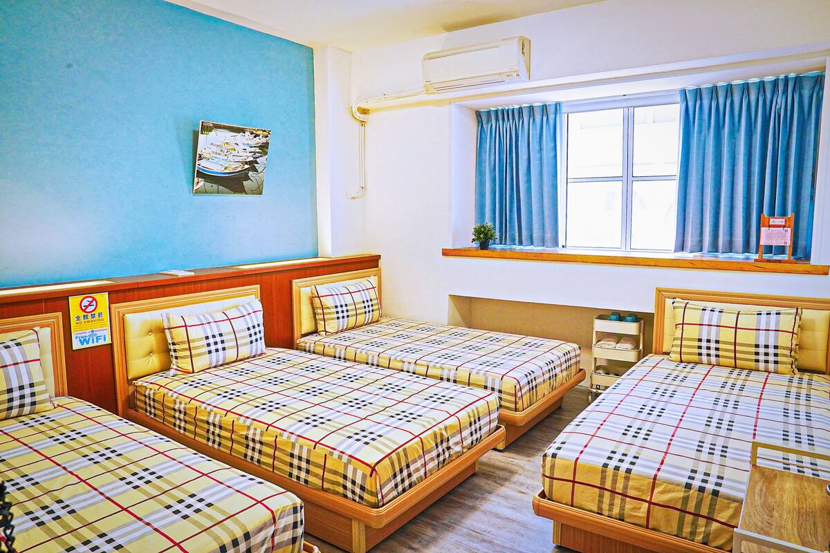 A hotel room with three beds, a window with blue curtains, a small plant on the windowsill, and a sign indicating WiFi availability.