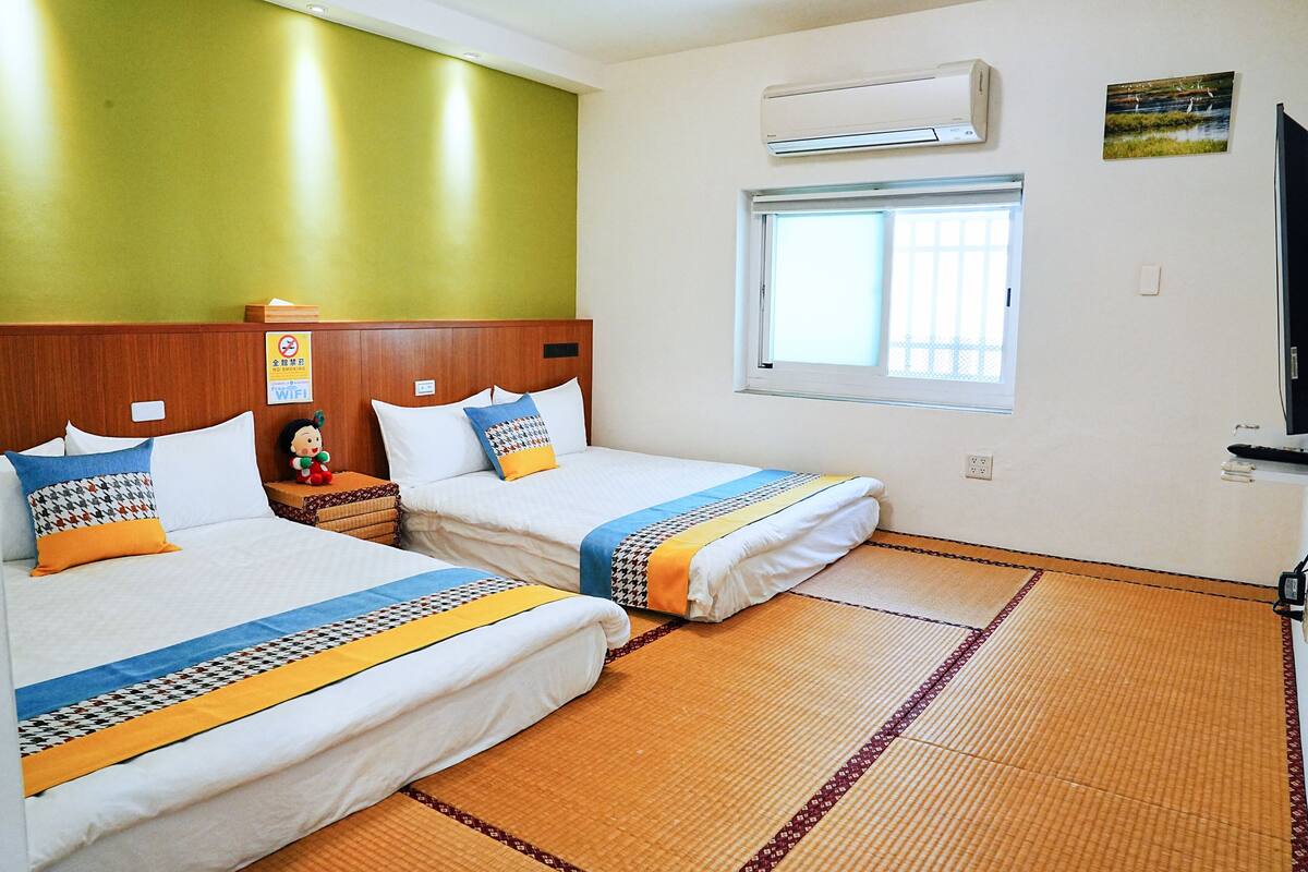 A hotel room with two beds, a wooden headboard, a window with blinds, an air conditioner, and a small table with a toy.