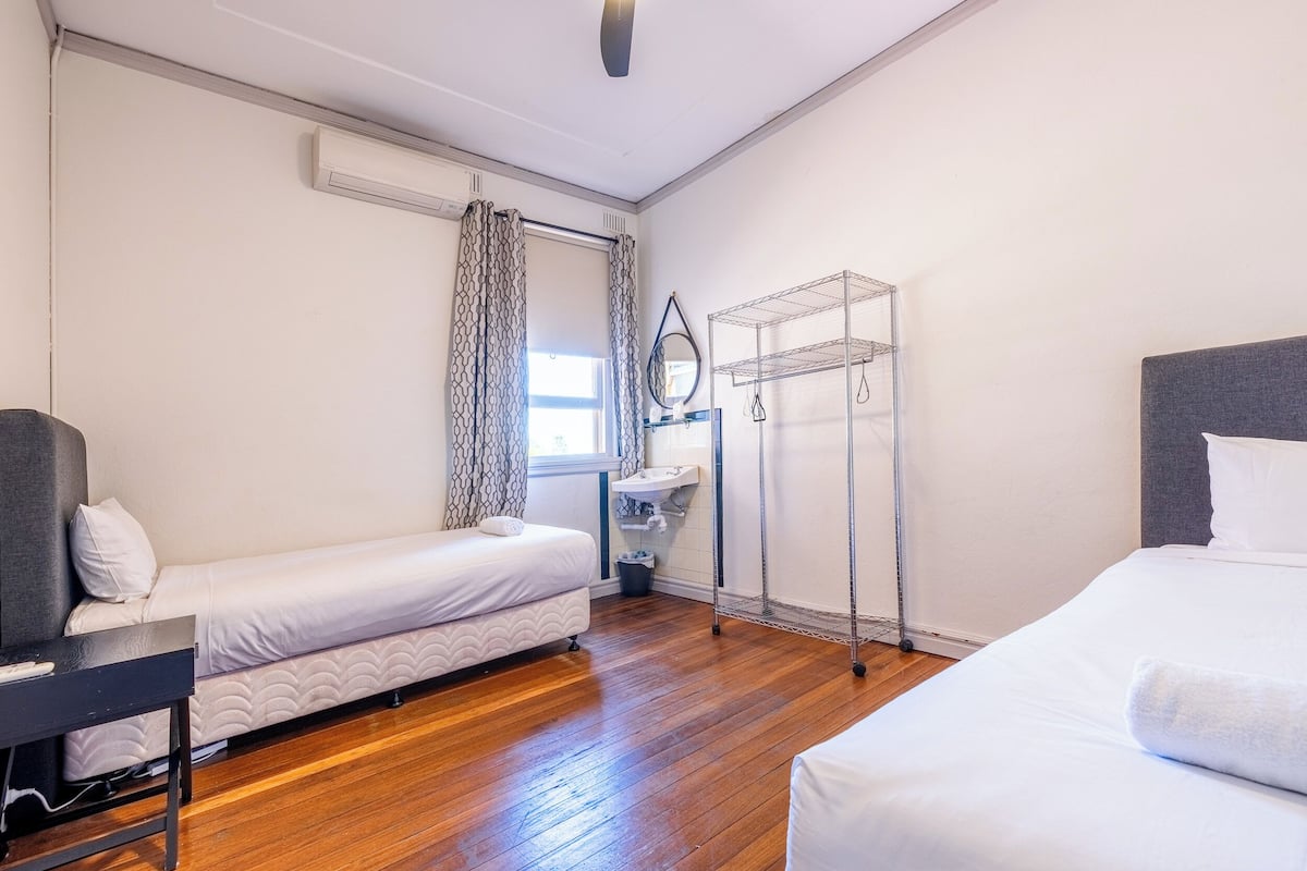 Ein Hotelzimmer mit Holzboden, zwei Einzelbetten, einem Schreibtisch, einem Spiegel, einem Wäscheständer aus Metall und einem Fenster mit Vorhängen.